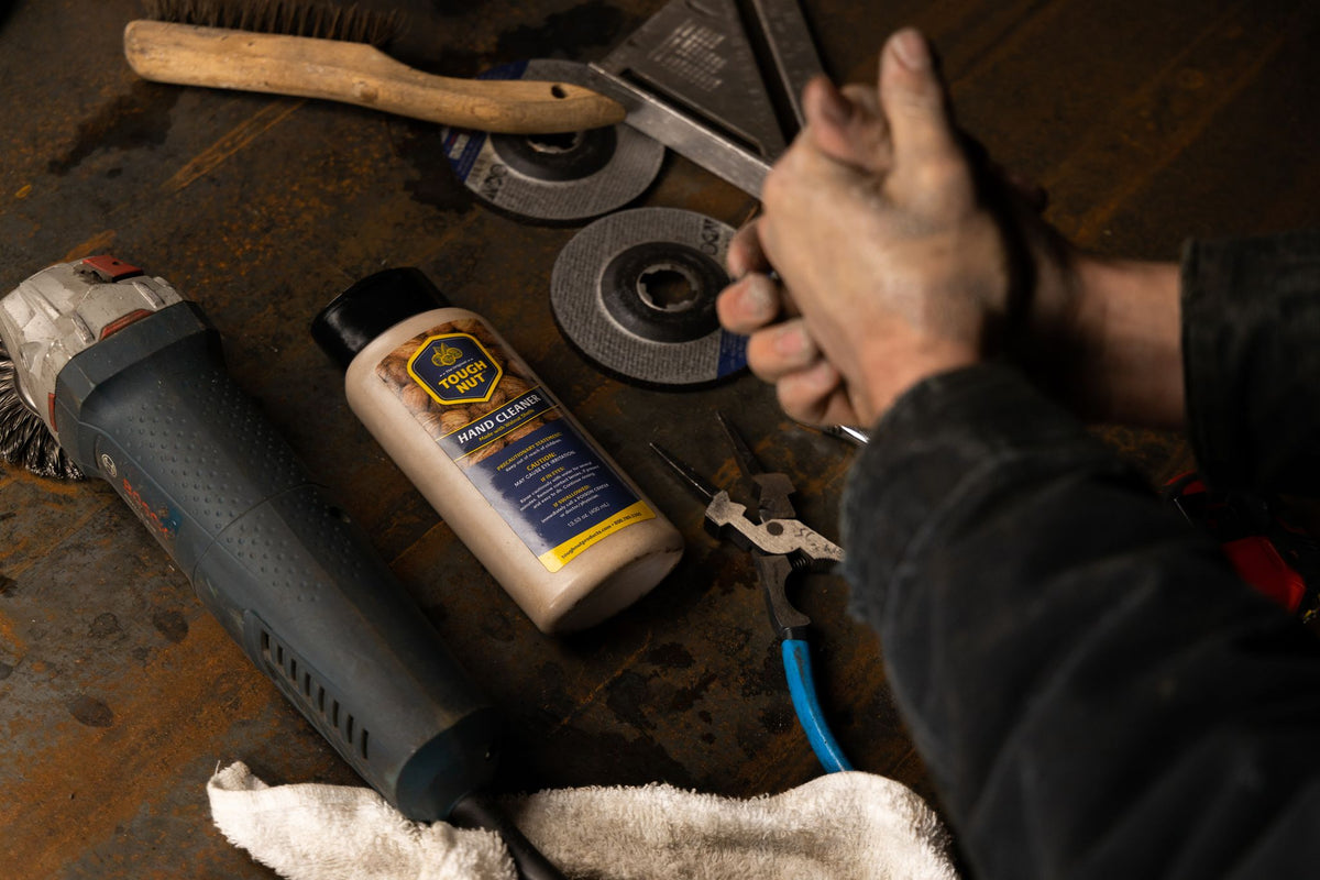 Top-down view of Tough Nut hand cleaner on a rusty workbench with an angle grinder, pliers and a dirty hand
